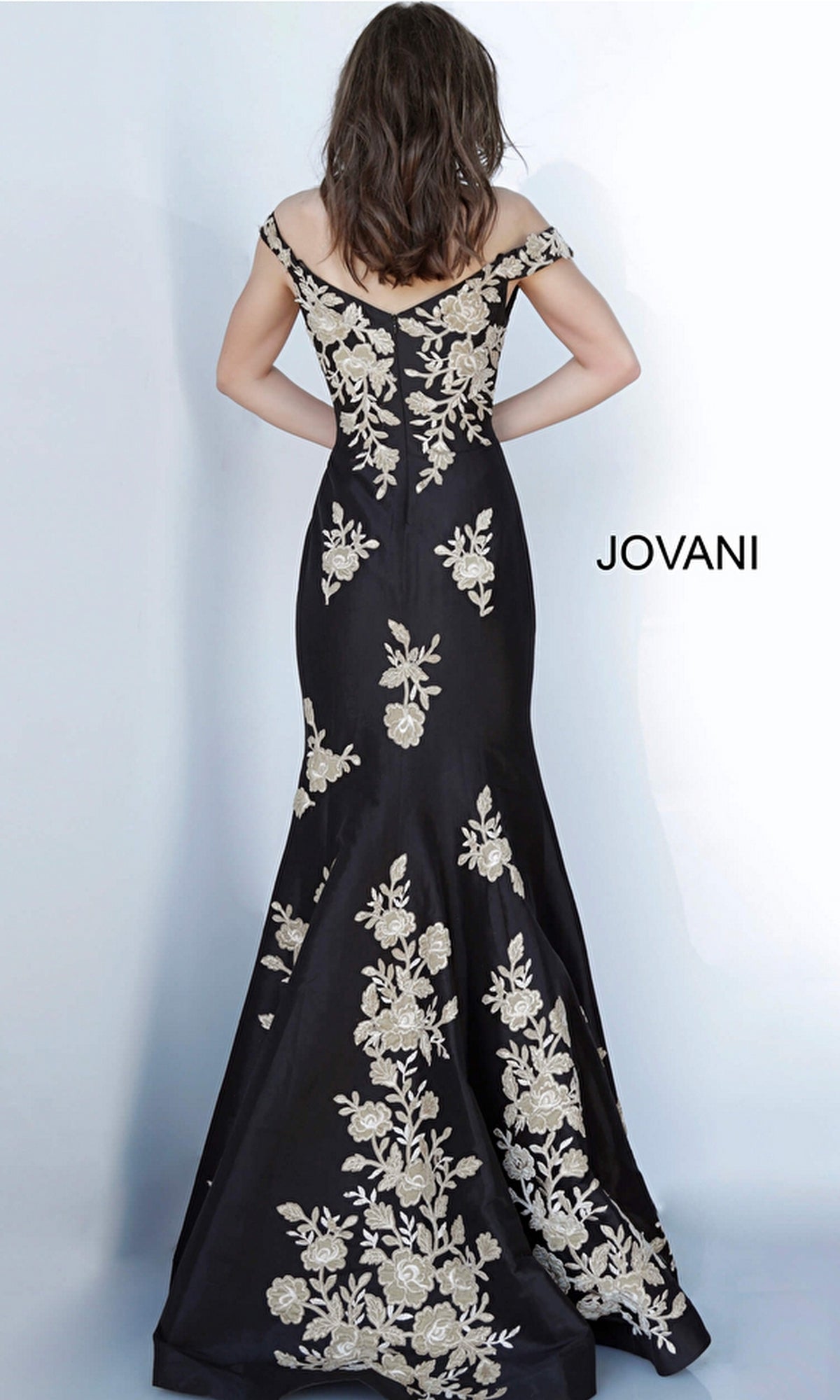 A woman faces away, wearing the Jovani Gold and Black Long Prom Dress 00635 with gold floral embroidery, against a plain light background.